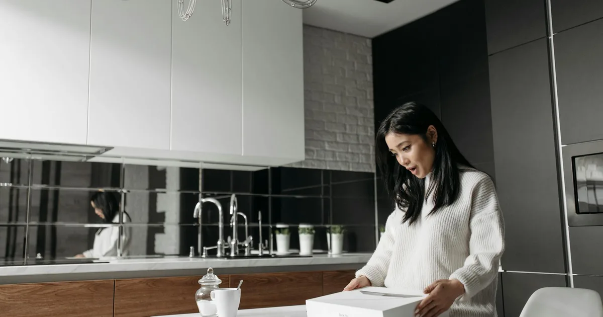 Asian woman in knitted sweater intrigued by package contents on kitchen table.