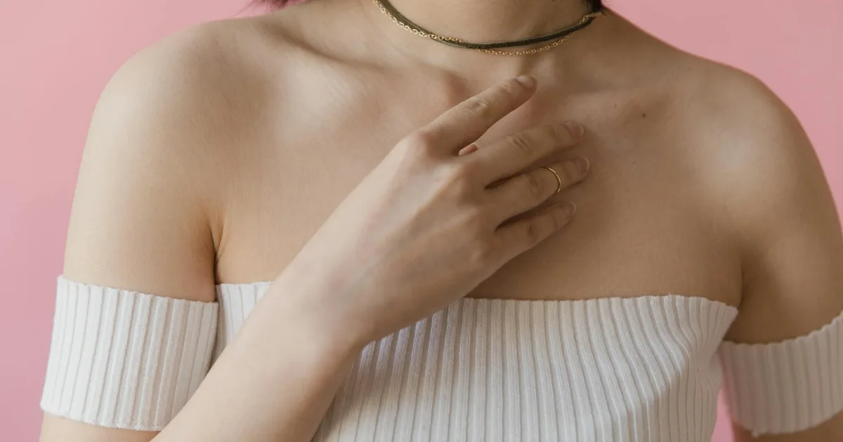 Close-up of a woman in a white off-shoulder top and necklace against a pink background.