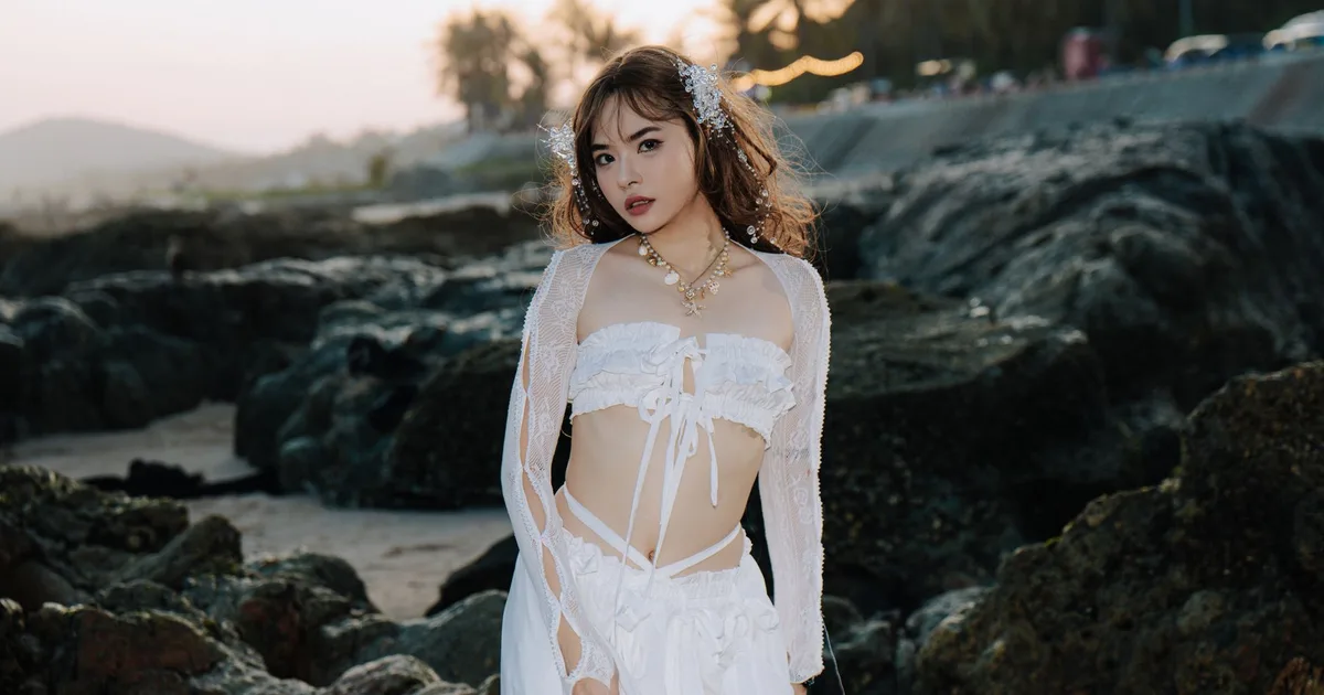 Stylish woman in a white dress poses by the rocky beach at sunset.