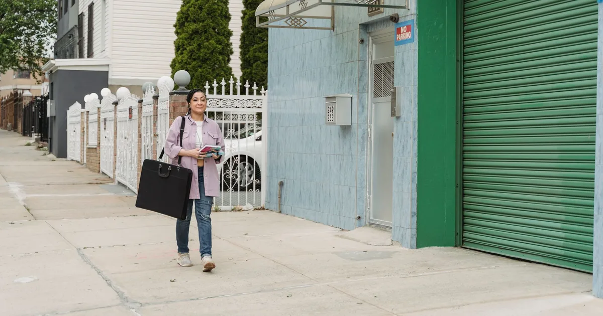 Full body of positive female with bag in casual clothes walking near buildings on pavement on street while smiling and looking forward