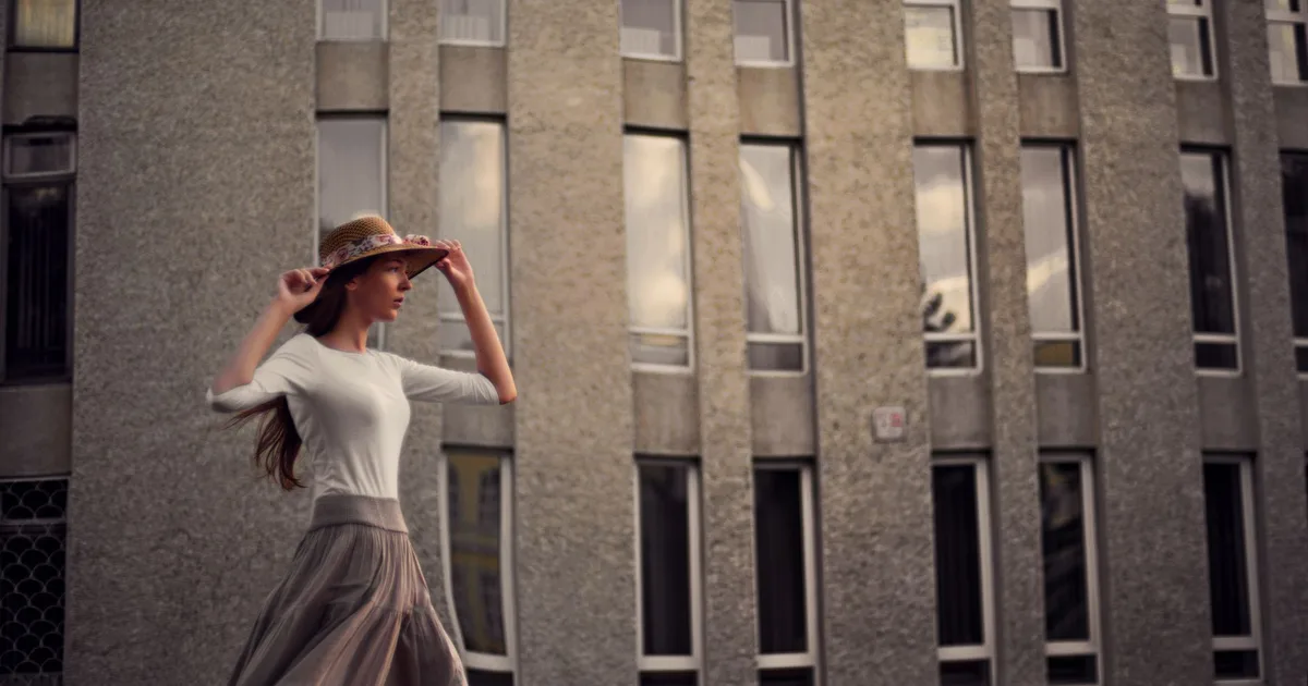 Stylish woman in a sun hat poses against a modern building facade on a windy day.