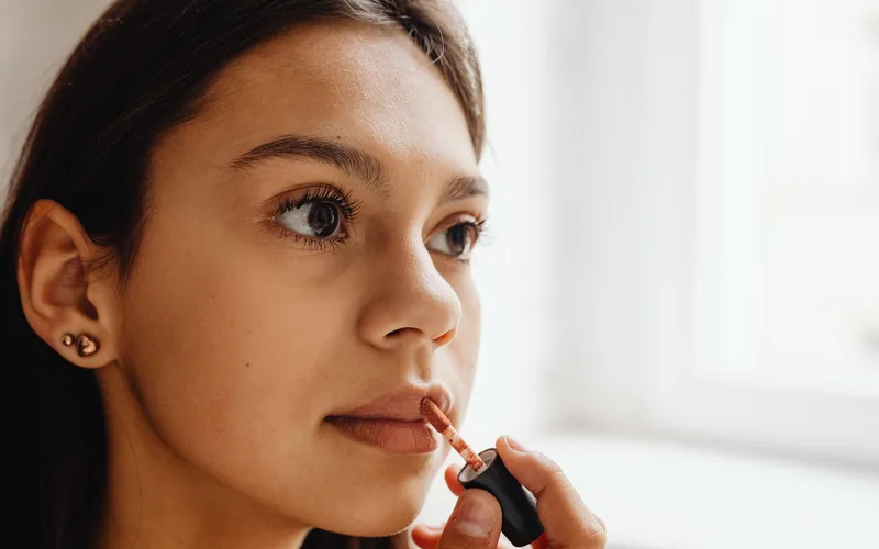 Close-up of a young woman applying lipstick indoors with natural daylight.