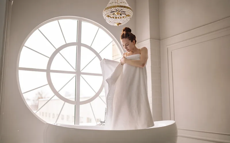 A woman wrapped in a towel stands elegantly in a bathtub by a large window.