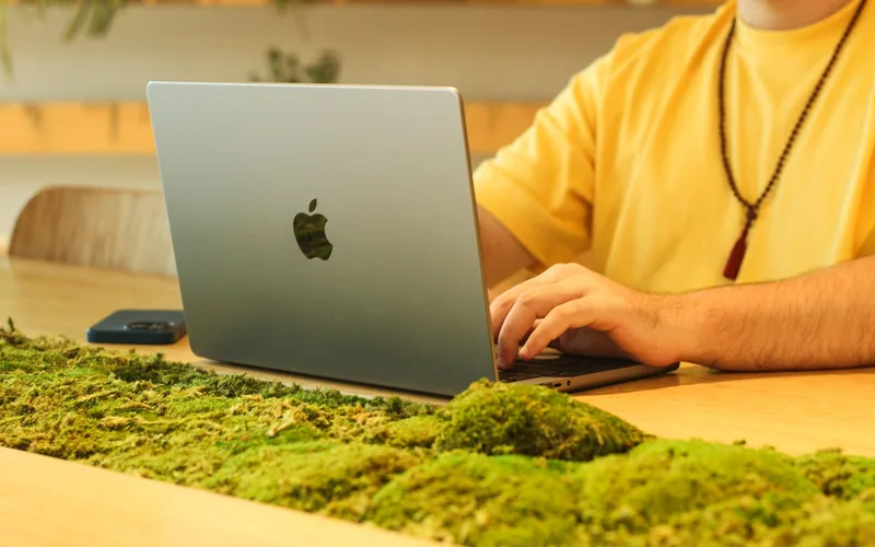 Man in yellow shirt working on laptop at a moss-decorated wooden table indoors.