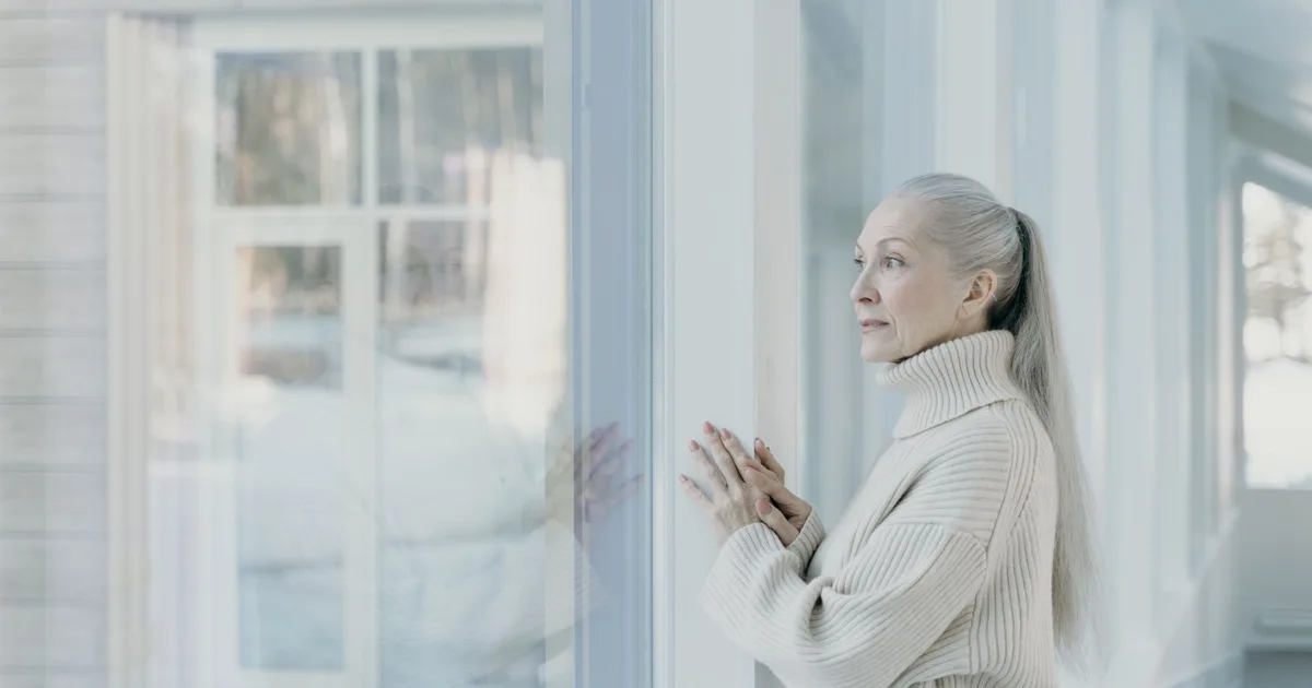 Serene elderly woman with grey hair looking out the window in a bright room, wearing a cozy sweater.