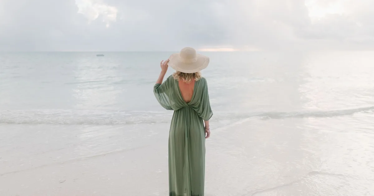 Back view of unrecognizable tender female wearing summer dress and straw hat standing on seashore and enjoying seascape during vacation