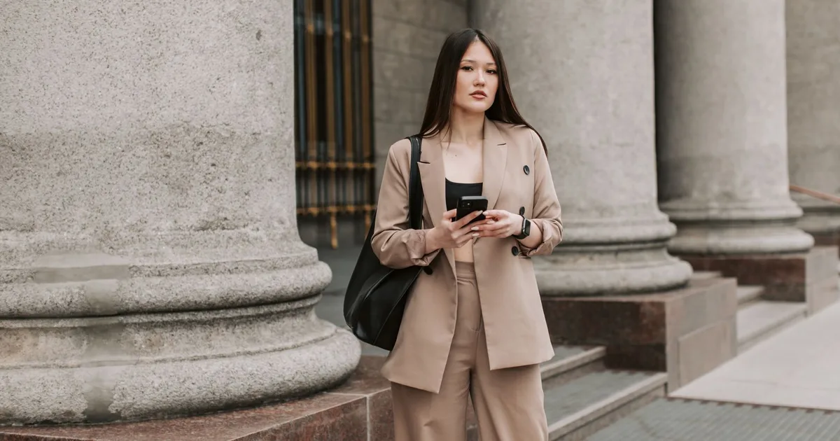 Young woman in beige business suit holding smartphone, standing near columns outside.