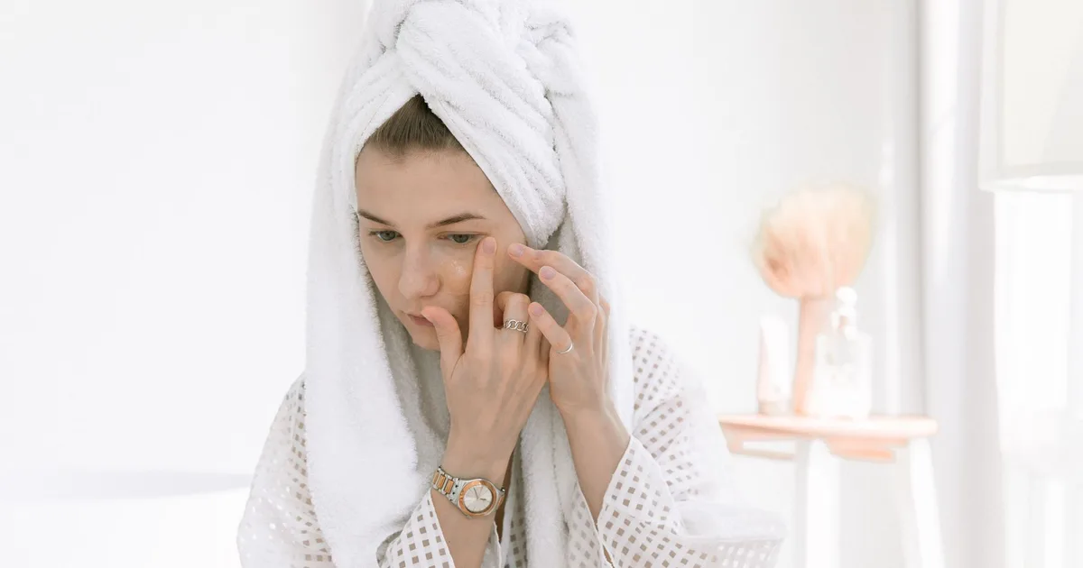 A woman in a bathrobe applies skincare cream in a bright, relaxing indoor setting.