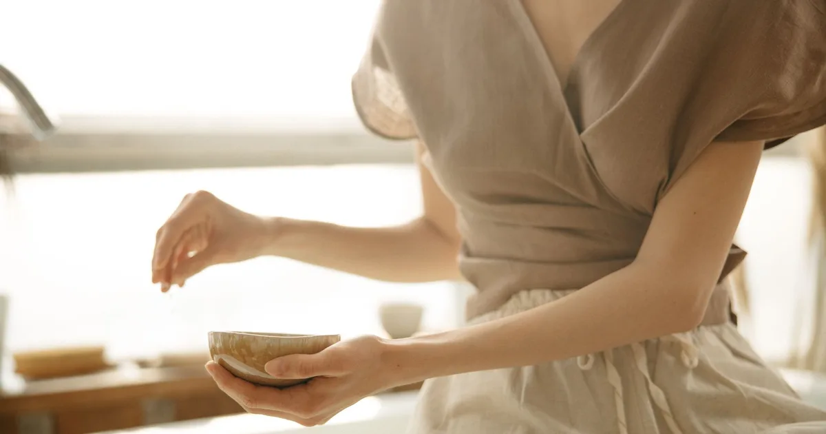 An unrecognizable woman sitting indoors, holding a bowl in soft natural light.