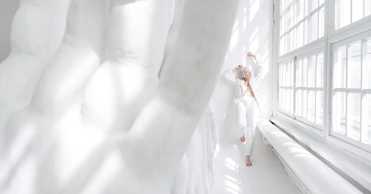 Minimalist fashion photo of a woman in white attire posing in a sunlit room with large windows.