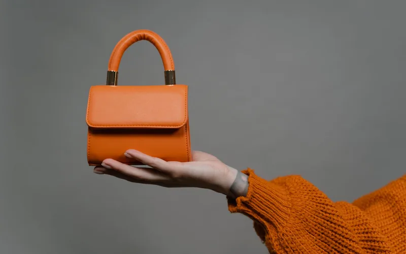 Stylish orange handbag held by a hand against a gray background. Perfect for modern fashion.