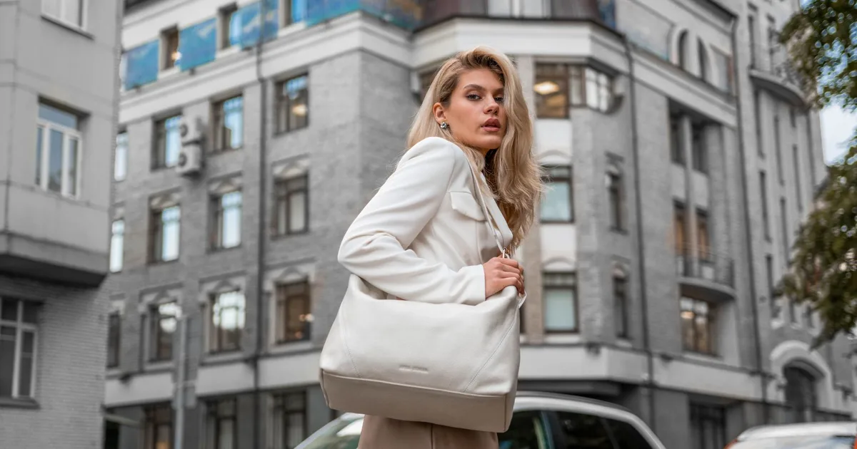 Stylish woman in white outfit and bag walking through city street.
