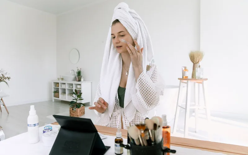 A woman in a bathrobe applies skincare products in a bright bathroom setting.