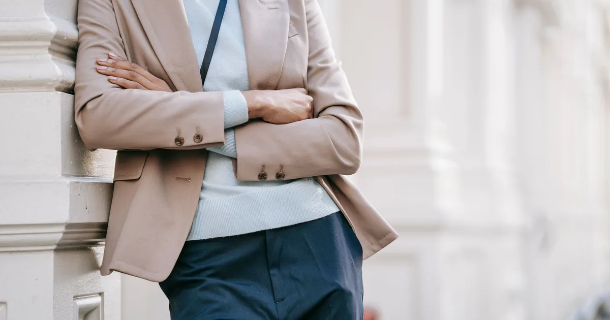 Crop anonymous stylish female manager in smart casual outfit leaning on elegant building wall with folded arms while standing on street on sunny day
