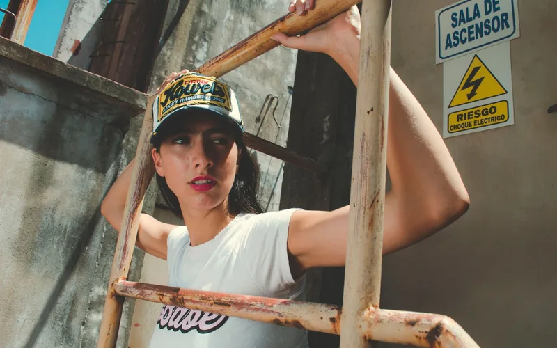 Model in urban setting holding a rusted handrail near a warning sign, emphasizing edgy fashion.
