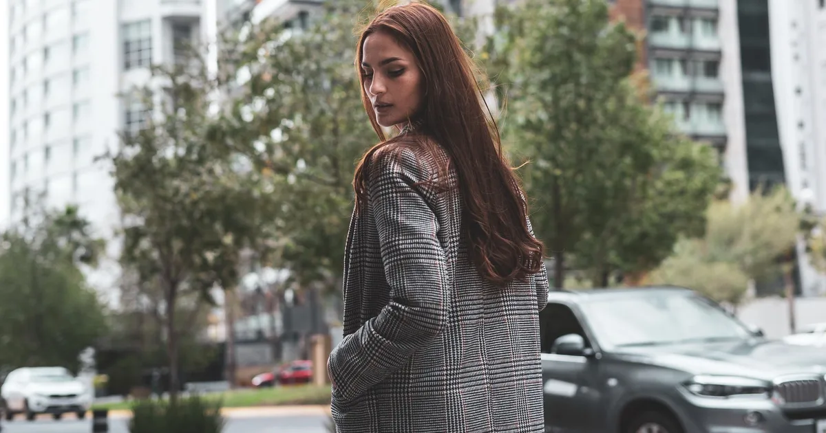 A fashionable woman in a checkered coat stands in a cityscape, embodying urban elegance.